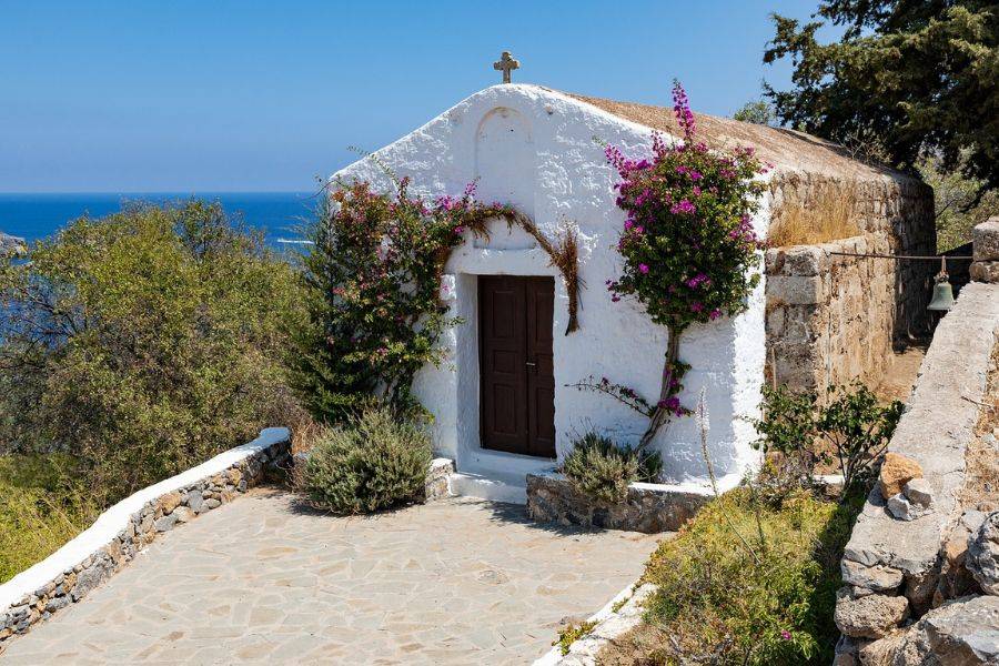 Eine kleine, traditionelle weiße Kapelle mit einer braunen Holztür, geschmückt mit rosa Bougainvillea-Blüten, thront auf einer Anhöhe mit Blick auf das blaue Meer.