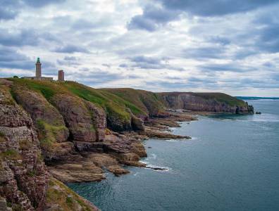 Blick auf die steilen Klippen des Cap Fréhel in der Bretagne mit dem markanten Leuchtturm und dramatisch bewölktem Himmel über dem Meer.