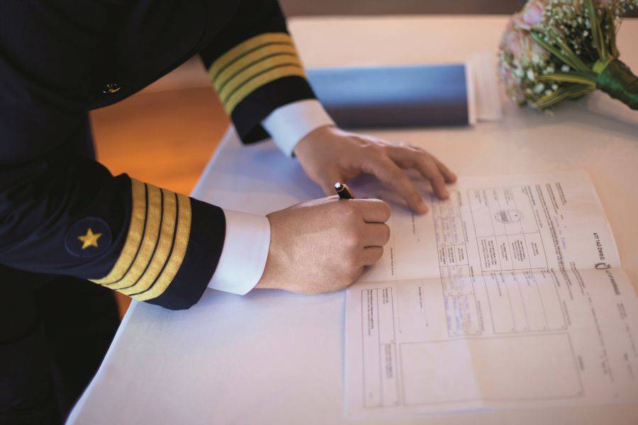 Kapitän in Uniform unterschreibt ein offizielles Dokument, vermutlich eine Heiratsurkunde, auf einem weißen Tisch mit einem Brautstrauß im Hintergrund.