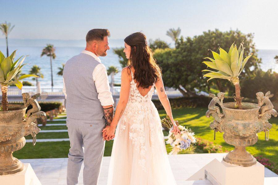 Brautpaar blickt Hand in Hand von einer Terrasse aufs Meer, umgeben von mediterranen Pflanzen und festlichem Ambiente.