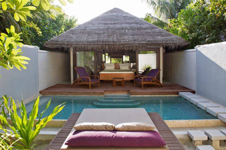 Gemütliche Poolvilla mit Strohdach, umgeben von tropischem Grün, mit Liegestühlen und Tagesbett auf Holzdeck vor dem Schlafzimmerbereich.