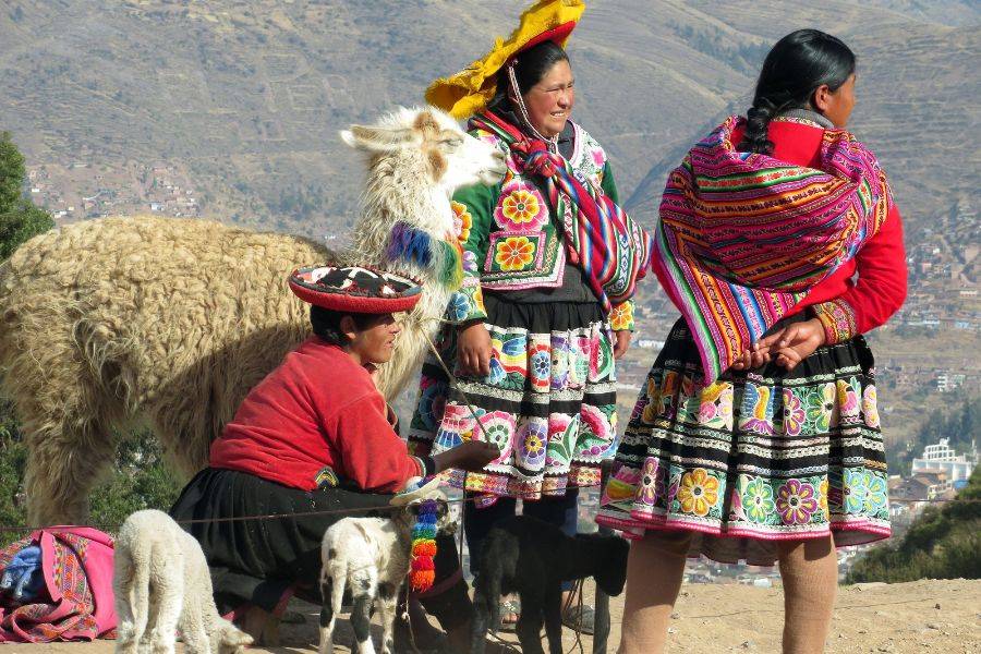 Drei Frauen in farbenfroher, traditioneller Kleidung der Anden posieren mit einem Alpaka und mehreren kleinen Tieren vor einer Berglandschaft.