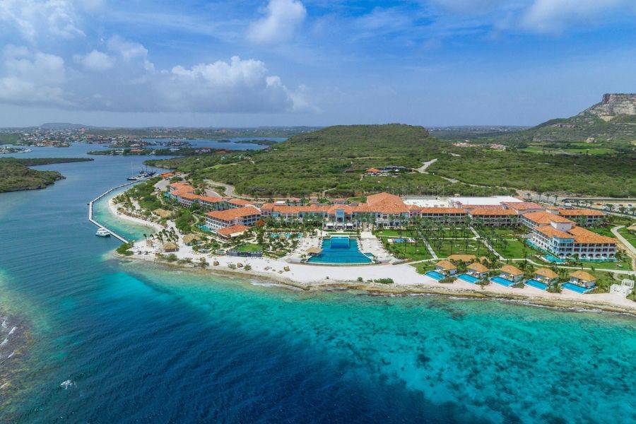  Luftaufnahme eines weitläufigen Luxusresorts mit roten Dächern direkt an der Küste, umgeben von türkisblauem Wasser und grüner Hügellandschaft.