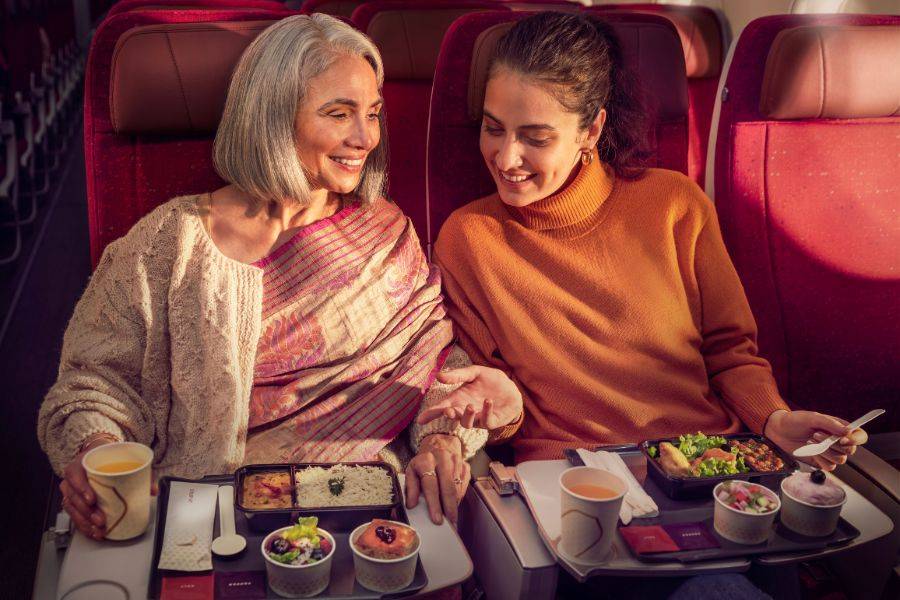 Zwei Passagierinnen genießen eine warme Mahlzeit an Bord von Air India in der Economy Class und lächeln einander an.