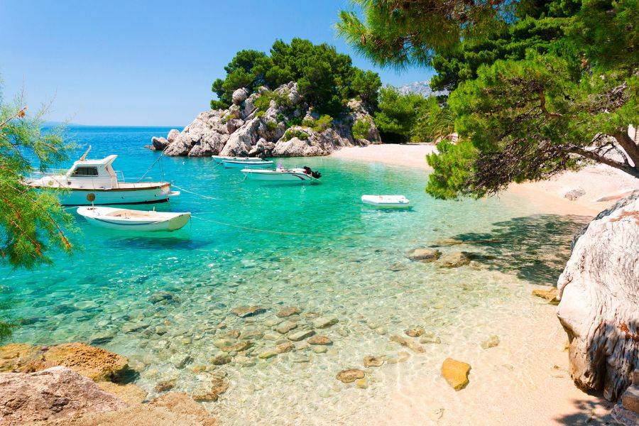 Kleine Boote liegen vor Anker in einer türkisfarbenen Bucht mit Sandstrand, Felsen und Pinien
