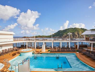 Ein luxuriöses Kreuzfahrtschiff mit einem großzügigen Poolbereich auf dem Sonnendeck. Der Pool ist von Liegestühlen umgeben, und im Hintergrund sieht man eine malerische Insel mit grünen Hügeln unter einem klaren, blauen Himmel mit wenigen Wolken. 