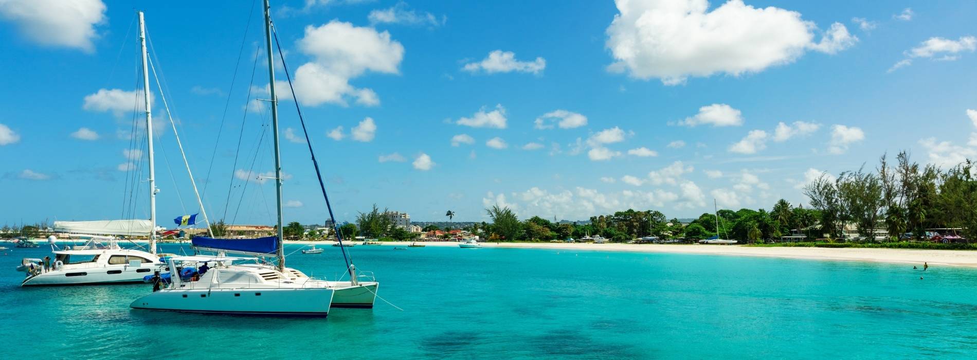 Segelboote und Katamarane liegen vor einem weißen Sandstrand im türkisblauen Karibikmeer unter strahlend blauem Himmel mit vereinzelten Wolken.
