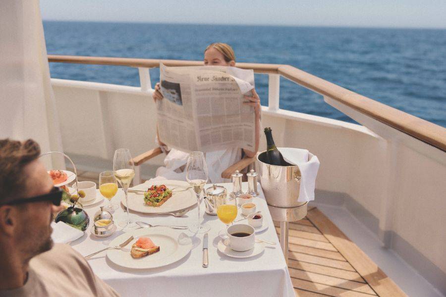Gäste genießen ein Frühstück an einem gedeckten Tisch an Deck eines Kreuzfahrtschiffs mit Blick auf das offene Meer