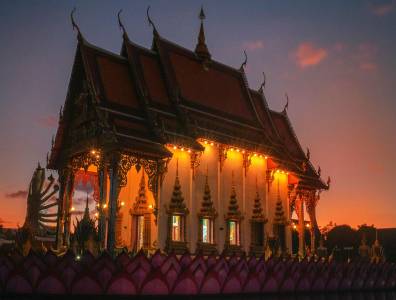 Ein buddhistischer Tempel bei Sonnenuntergang, beleuchtet von warmen Lichtern, mit kunstvollen, traditionellen Verzierungen und einem Himmel in sanften Rot- und Orangetönen im Hintergrund.