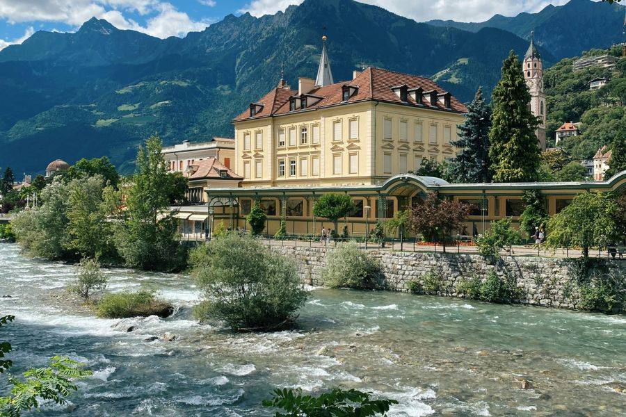 Blick auf das historische Kurhaus in Meran, direkt am klaren Passerfluss gelegen, mit Bergen im Hintergrund und blühendem Grün entlang des Ufers.