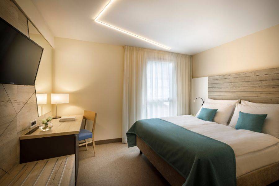 Das Bild zeigt ein modernes und minimalistisches Hotelzimmer. Das Doppelbett ist mit weißen Bettlaken und türkisfarbenen Kissen sowie einer passenden Tagesdecke ausgestattet. 