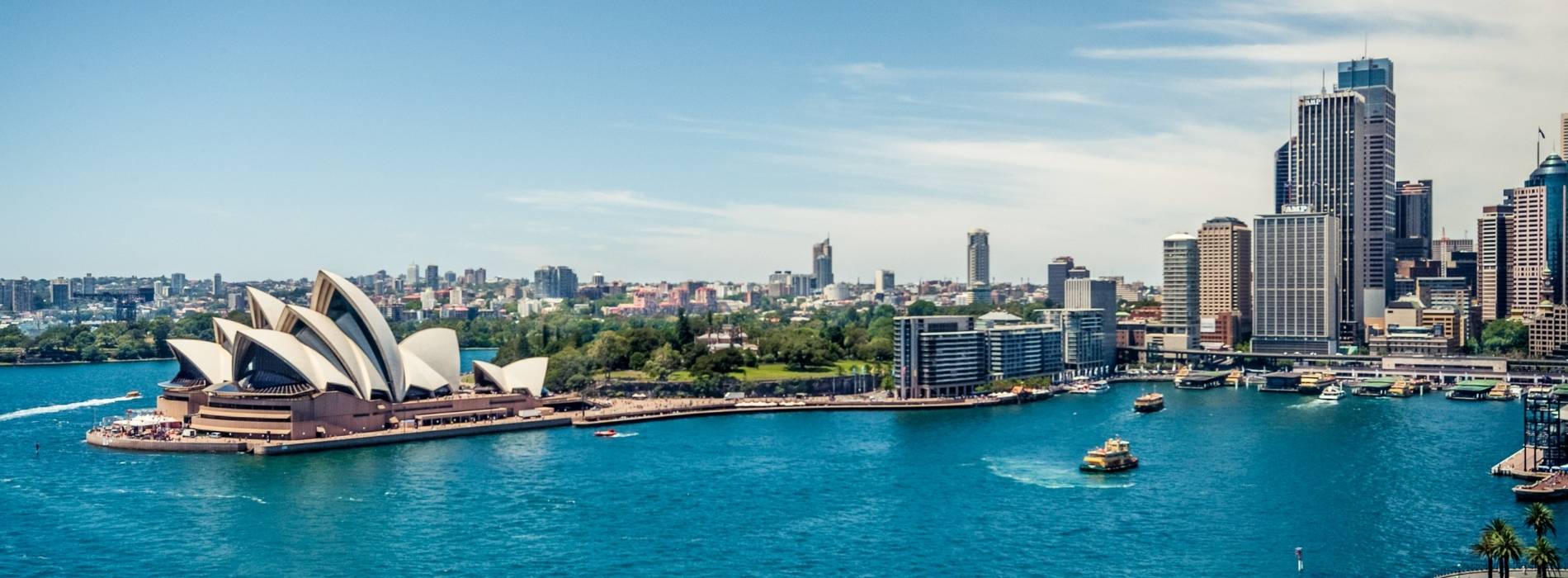 Ein Panoramablick auf den Hafen von Sydney in Australien. Das Sydney Opera House ist deutlich links zu sehen, mit seinen ikonischen weißen, segelförmigen Dächern. Das Wasser des Hafens ist hellblau, und mehrere Boote fahren hindurch. Im Hintergrund ist die Stadtsilhouette mit modernen Hochhäusern unter klarem Himmel sichtbar.