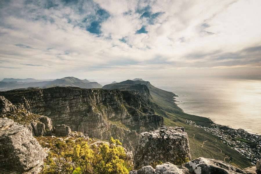 Eine dramatische und weite Aussicht vom Gipfel des Tafelbergs, mit Blick auf felsige Klippen und das Meer. Die Landschaft erstreckt sich bis zum Horizont, mit Wolken, die darüber schweben.
