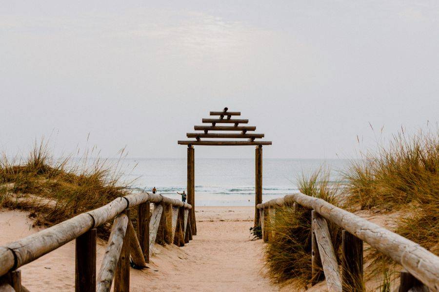 Dieses Bild fängt die ruhige Schönheit der Costa de la Luz in Andalusien ein. Der Holzsteg führt durch sanfte Dünen hinunter zum weiten Sandstrand, wo das Meer in sanften Wellen ans Ufer rollt. 