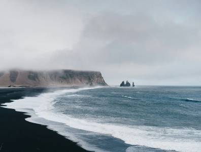 Weitläufiger schwarzer Sandstrand in Island mit rauem Meer, Felsformationen und nebliger Küstenlandschaft.