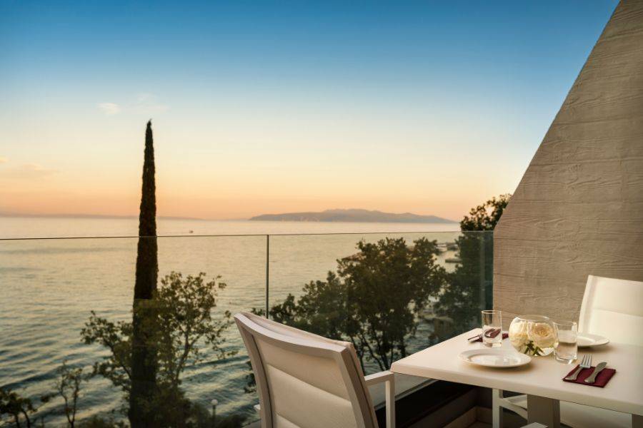 Romantische Terrasse mit einem gedeckten Tisch für zwei, Glasgeländer und Blick auf das ruhige Meer und einen warmen Sonnenuntergang am Horizont.