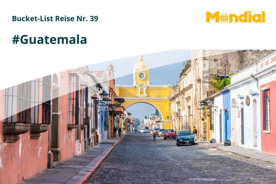 Bunte Kolonialhäuser und Kopfsteinpflasterstraße in der Altstadt von Antigua, Guatemala, mit dem berühmten gelben Santa-Catalina-Bogen im Vordergrund und einem Vulkan im Hintergrund; Reisekampagne von Mondial mit dem Hashtag #Guatemala und dem Titel „Bucket-List Reise Nr. 39“