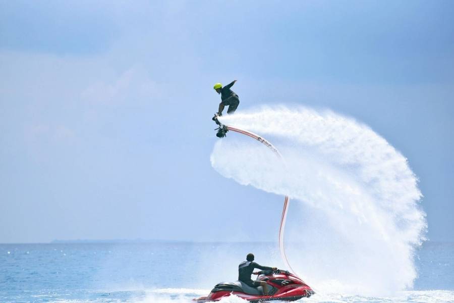 Person auf einem Flyboard hoch über dem Meer, getragen von Wasserstrahlen, mit einem Jetski darunter.