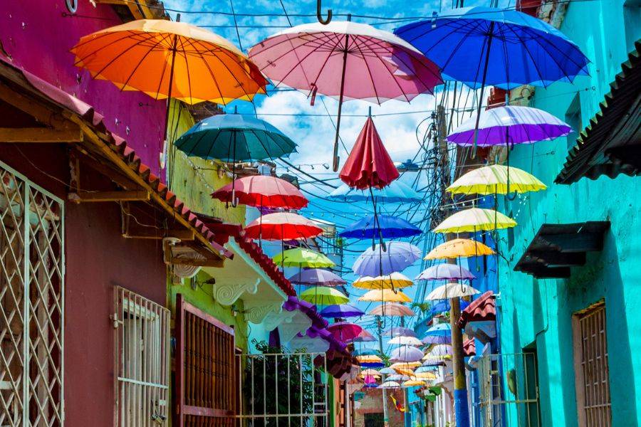 © VERKEHRSBUERO Bunte Gasse in Cartagena, Kolumbien, mit schwebenden Regenschirmen als Dekoration.