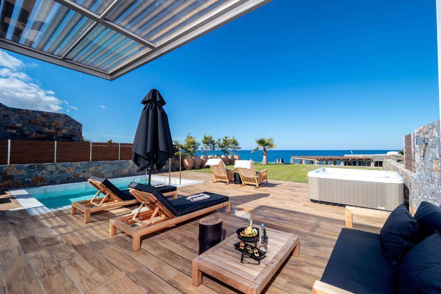 Luxuriöse Holzterrasse mit Sonnenliegen, Whirlpool und Pool, mit Blick auf das Meer unter strahlend blauem Himmel.