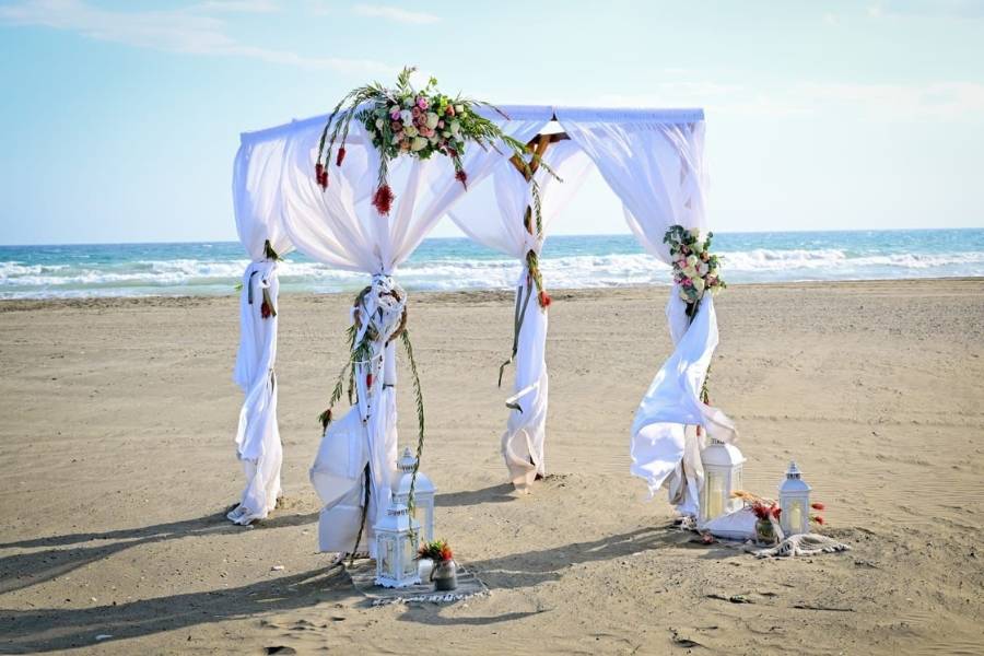 Romantischer Hochzeitsbogen am Strand von Zypern, dekoriert mit weißen Stoffbahnen, Blumenarrangements und Laternen – perfekte Kulisse für eine Strandhochzeit im Ausland.