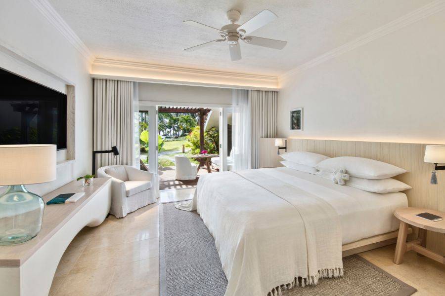 Lichtdurchflutetes Hotelzimmer in hellen Naturtönen mit großem Bett, Deckenventilator, modernem Interieur und Zugang zur Terrasse mit Blick ins Grüne.