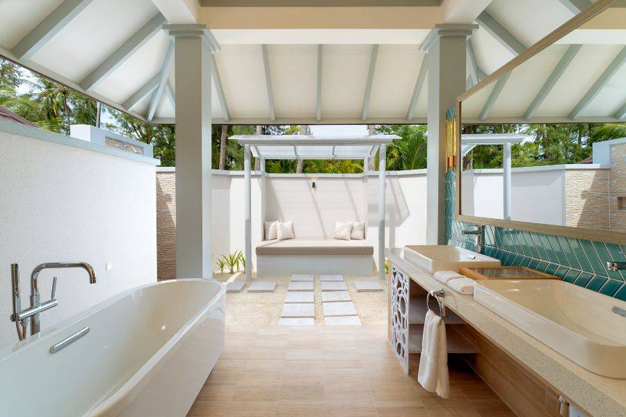 Elegantes, halb offenes Badezimmer mit freistehender Badewanne, Doppelwaschtisch und Lounge-Bereich im Freien, umgeben von tropischer Vegetation.