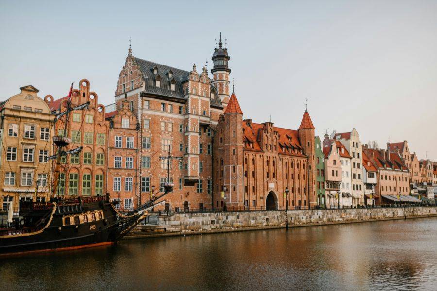 Die prächtige Uferpromenade der Altstadt mit dem berühmten Krantor (Żuraw) im Hintergrund – eines der Wahrzeichen von Danzig. Die historischen Backsteinhäuser und das stilisierte Piratenschiff sind typische Fotomotive am Fluss Motława.
