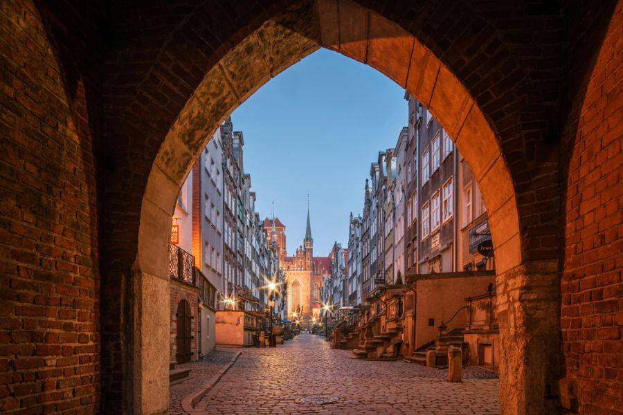 Ein stimmungsvoller Blick durch ein gotisches Tor in die Altstadtgasse mit dem bekannten Langen Markt (Długi Targ) im Hintergrund, wo sich das Rathaus und der Neptunbrunnen befinden.