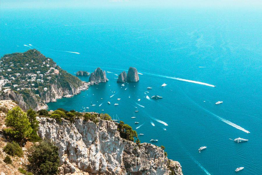 Blick auf die Insel Capri mit den berühmten Faraglioni-Felsen und zahlreichen Booten im tiefblauen Meer, aus luftiger Perspektive aufgenommen.