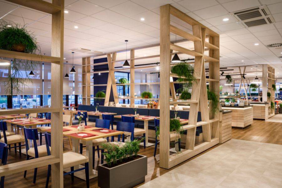 Modernes Restaurant mit kreativem Holzregal-Design, das Bereiche trennt, bunten Stühlen, stilvollen Tischdekorationen und grünen Pflanzen für eine lebendige Atmosphäre