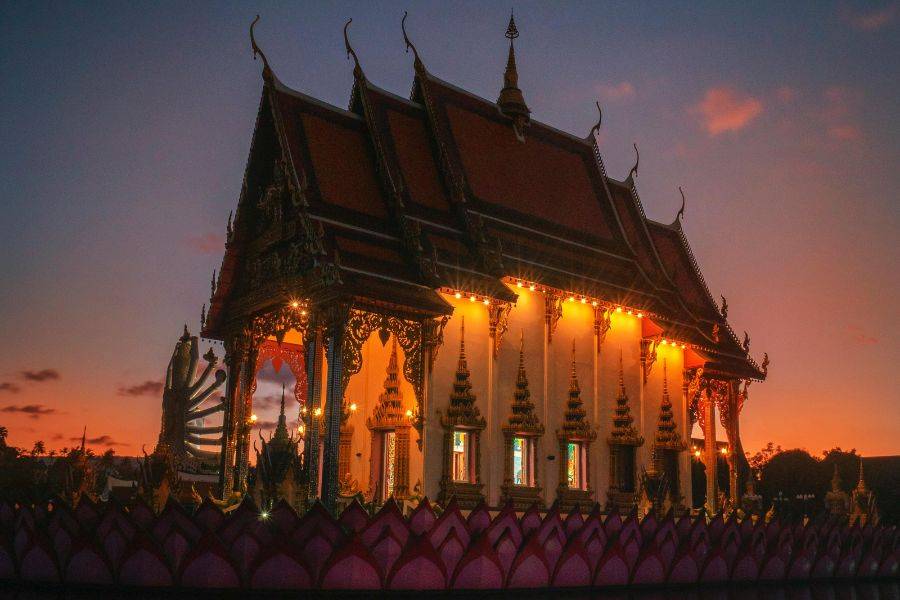 Ein buddhistischer Tempel bei Sonnenuntergang, beleuchtet von warmen Lichtern, mit kunstvollen, traditionellen Verzierungen und einem Himmel in sanften Rot- und Orangetönen im Hintergrund.
