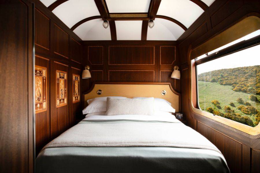 Ein stilvoll eingerichtetes Zugabteil mit dunkler Holzvertäfelung und kunstvollen Intarsien. Ein großes, komfortables Bett mit weichen Kissen und einer cremefarbenen Decke dominiert den Raum.