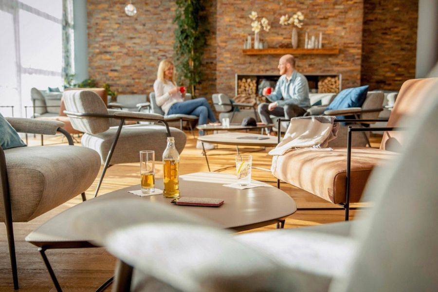 Das Bild zeigt eine moderne, gemütliche Lounge mit stilvollen Sitzmöbeln und einem einladenden Kamin im Hintergrund. Die warme Atmosphäre wird durch natürliche Materialien wie Holz und Stein unterstrichen.