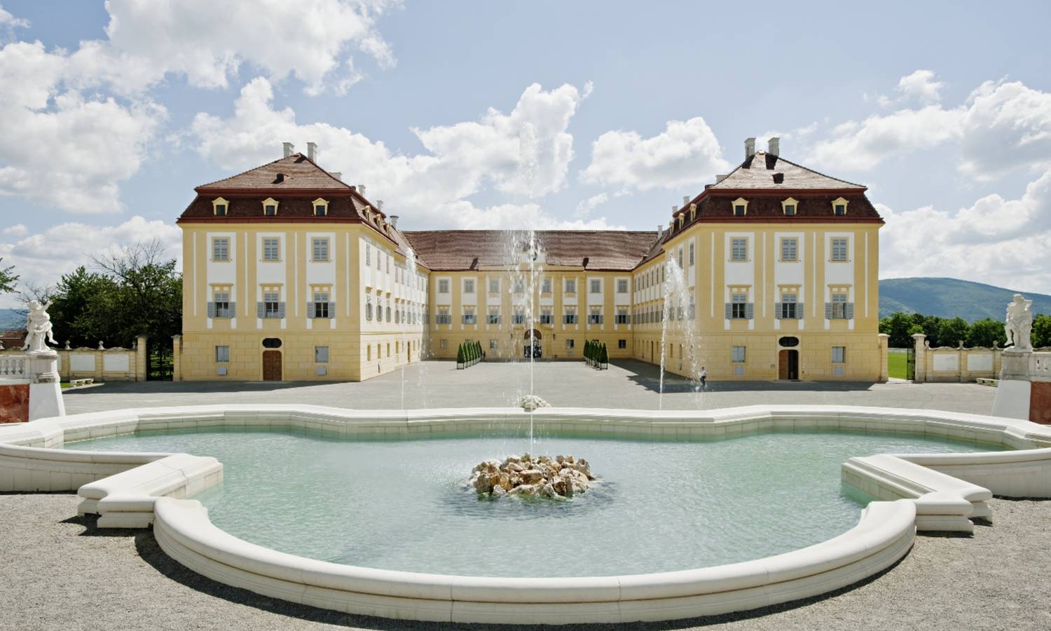 Ein prächtiges Schloss mit gelber Fassade, symmetrischen Flügeln und einem großen Springbrunnen im Vordergrund.