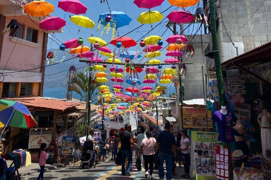 © Privat Bunte Regenschirme hängen dekorativ über einer lebhaften Marktstraße im guatemaltekischen Ort San Juan La Laguna am Atitlán-See; Menschen schlendern zwischen kleinen Geschäften, Essensständen und Souvenirläden bei strahlendem Sonnenschein.