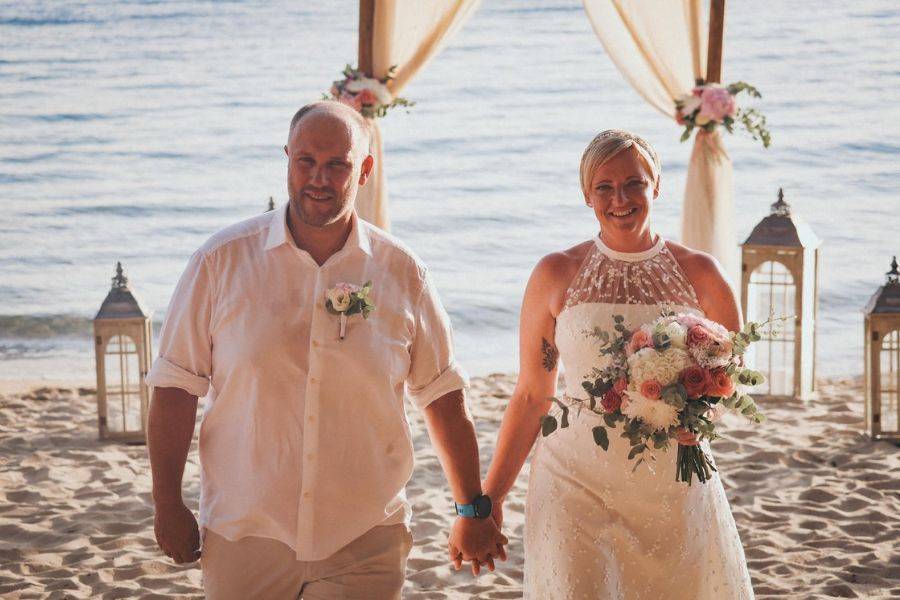 Hochzeitspaar geht Hand in Hand am Strand entlang, festlich gekleidet unter einem romantisch dekorierten Pavillon mit Blick aufs Meer.