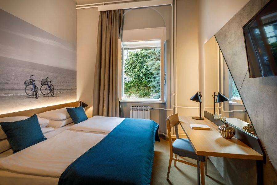 Modernes Hotelzimmer mit zwei Einzelbetten, minimalistischer Einrichtung, sanfter Beleuchtung und einem Fenster mit Blick auf grüne Bäume und ein Gebäude im Hintergrund.