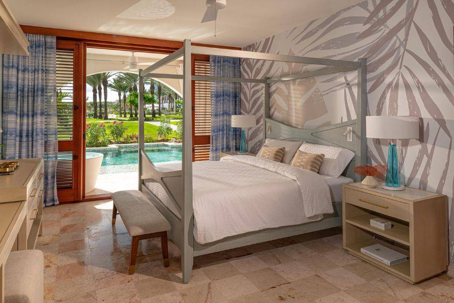 Modernes Hotelzimmer mit Himmelbett und direktem Zugang zur Terrasse mit Pool, mit Blick auf gepflegte Gartenanlage und Palmen.
