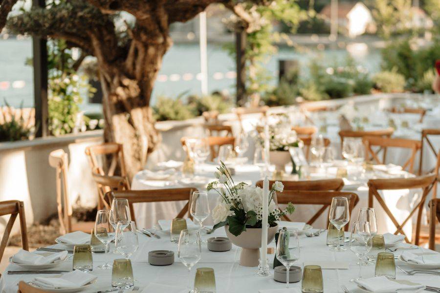 Elegant gedeckte Tische mit weißen Tischdecken und Blumenschmuck unter Bäumen in einem Garten mit Blick auf einen See, vorbereitet für eine Hochzeitsfeier bei Tageslicht.