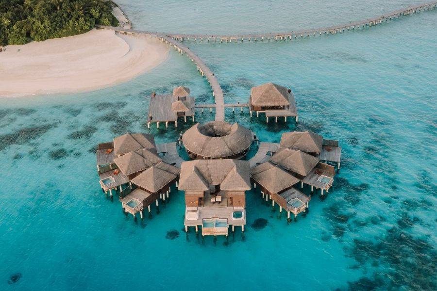 Luftaufnahme eines exklusiven Wasserresorts mit mehreren strohgedeckten Bungalows auf Stelzen, die kreisförmig über türkisfarbenem Meer angeordnet sind, mit Steg zur Insel.