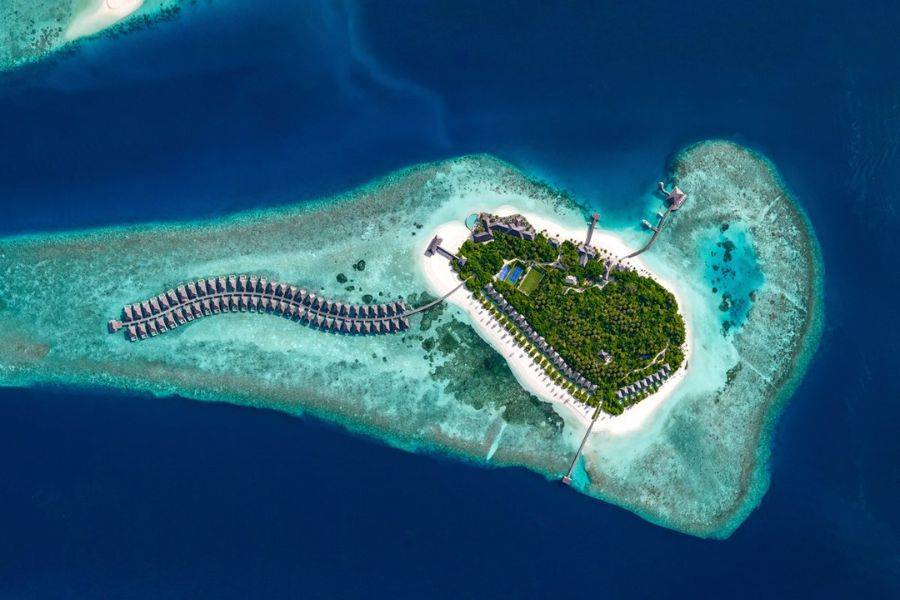 Eine malerische Luftaufnahme eines tropischen Inselresorts, umgeben von türkisfarbenem Wasser. Die Insel ist üppig bewachsen und verfügt über einen langen Steg mit Überwasservillen, die sich sanft in die Lagune einfügen. 
