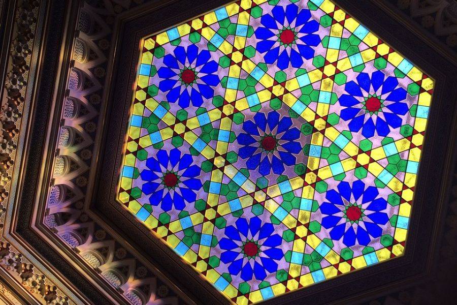 Buntes Mosaik aus Glas in geometrischen Mustern, mit dominierenden Blau-, Grün- und Gelbtönen in einer kunstvollen Deckenverzierung.