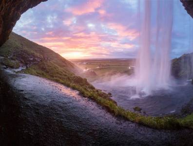 Blick hinter einen Wasserfall in Island bei Sonnenuntergang mit weichem Licht, grüner Landschaft und weiter Ebene im Hintergrund.