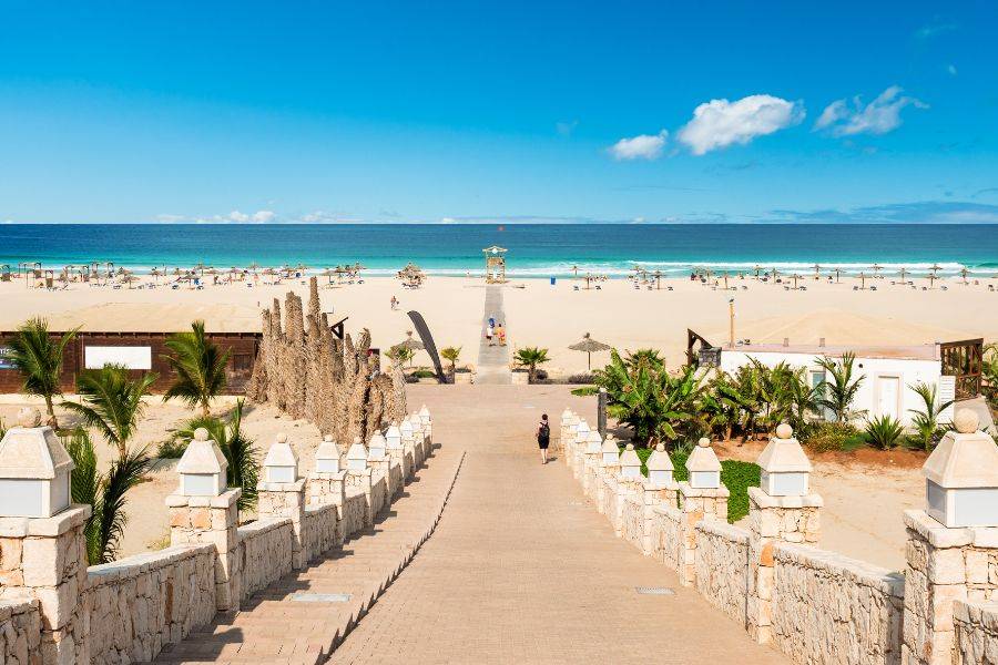Breiter Sandstrand mit Sonnenliegen und türkisfarbenem Meer, erreichbar über eine gepflegte Promenade mit Palmen und Steinmauer.