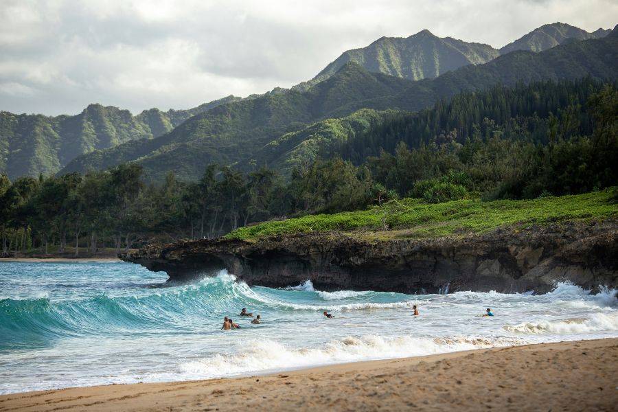 Wellen am naturbelassenen Strand der North Shore auf Oahu, Hawaii – grüne Berghänge, felsige Küste und Surfer im Wasser.