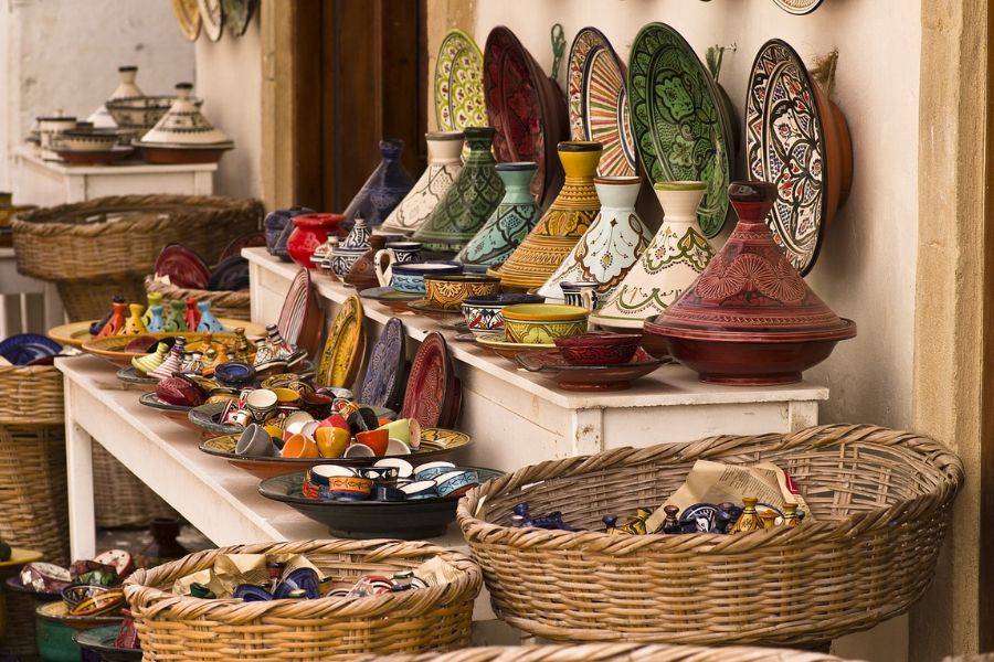 Ein Marktstand mit traditioneller marokkanischer Keramik, darunter farbenfrohe Tajine-Töpfe, Schalen und Teller, kunstvoll verziert mit orientalischen Mustern. Die Waren sind auf weißen Holztischen ausgestellt, umgeben von großen geflochtenen Körben.