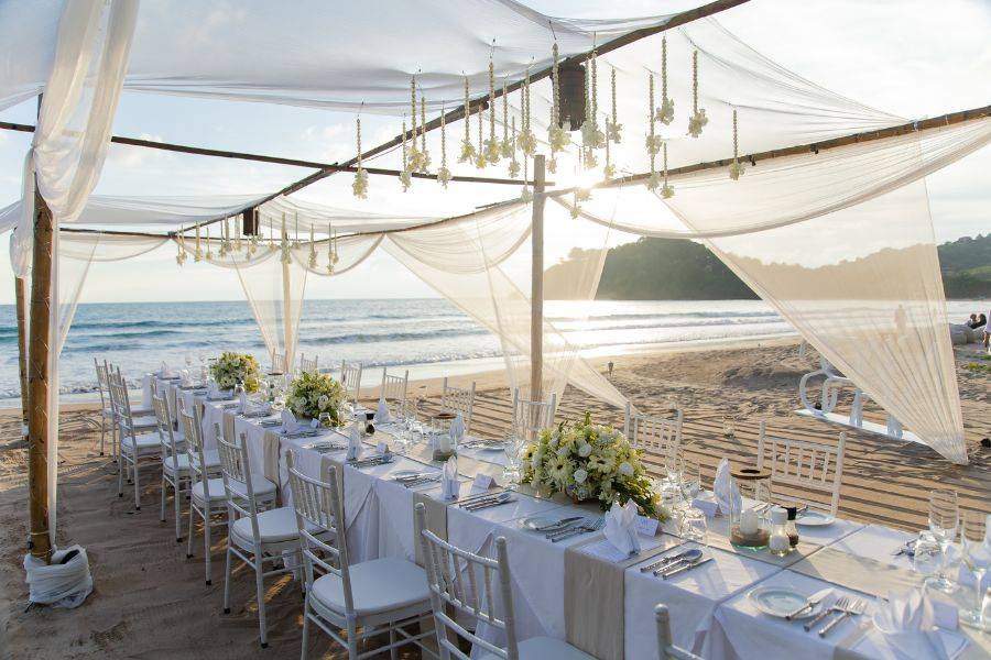 Festlich gedeckte lange Tafel am Strand unter einem transparenten, mit Blumen geschmückten Baldachin, mit Blick aufs Meer bei Sonnenuntergang.