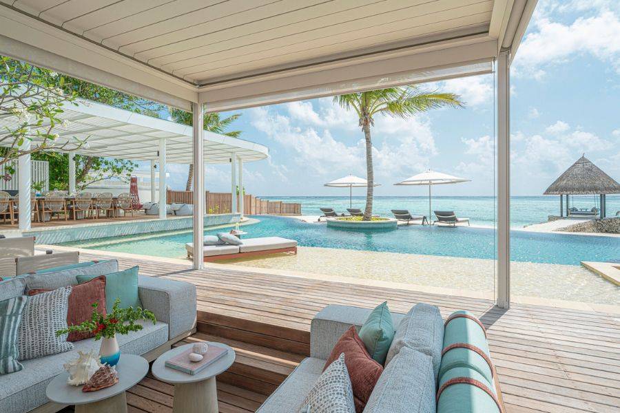 Offene Lounge einer stilvollen Strandvilla mit direktem Zugang zum Pool, eleganter Einrichtung und Aussicht auf das Meer mit Sonnenschirmen und Palmen.
