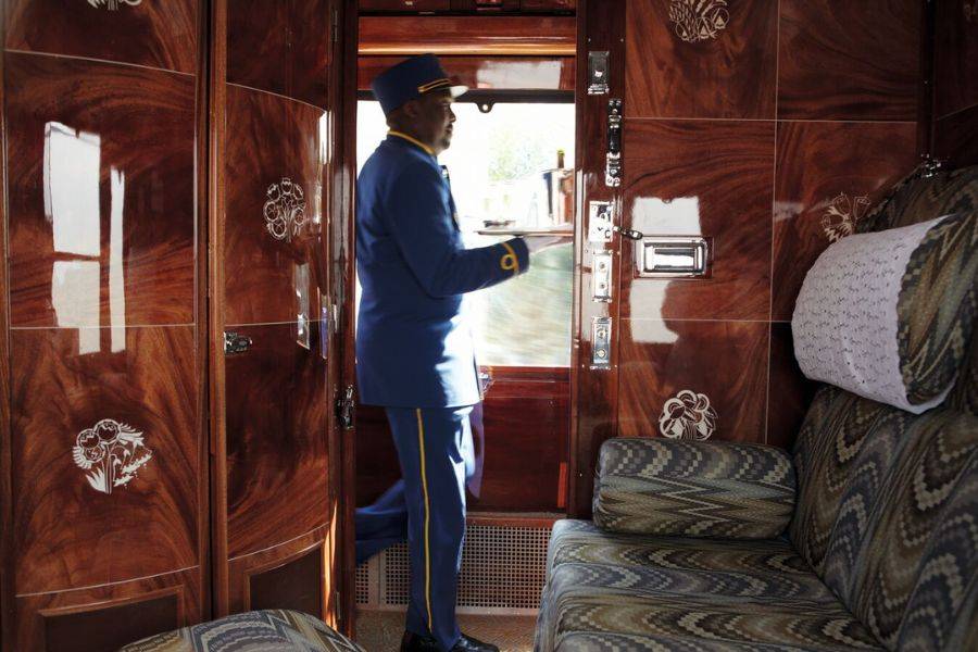 Ein Zugbegleiter in einer eleganten blauen Uniform mit goldenen Verzierungen bewegt sich durch ein luxuriöses Zugabteil. Er trägt ein Tablett mit Speisen oder Getränken, während er durch eine Tür mit glänzendem Holzfurnier und floralen Intarsien tritt.